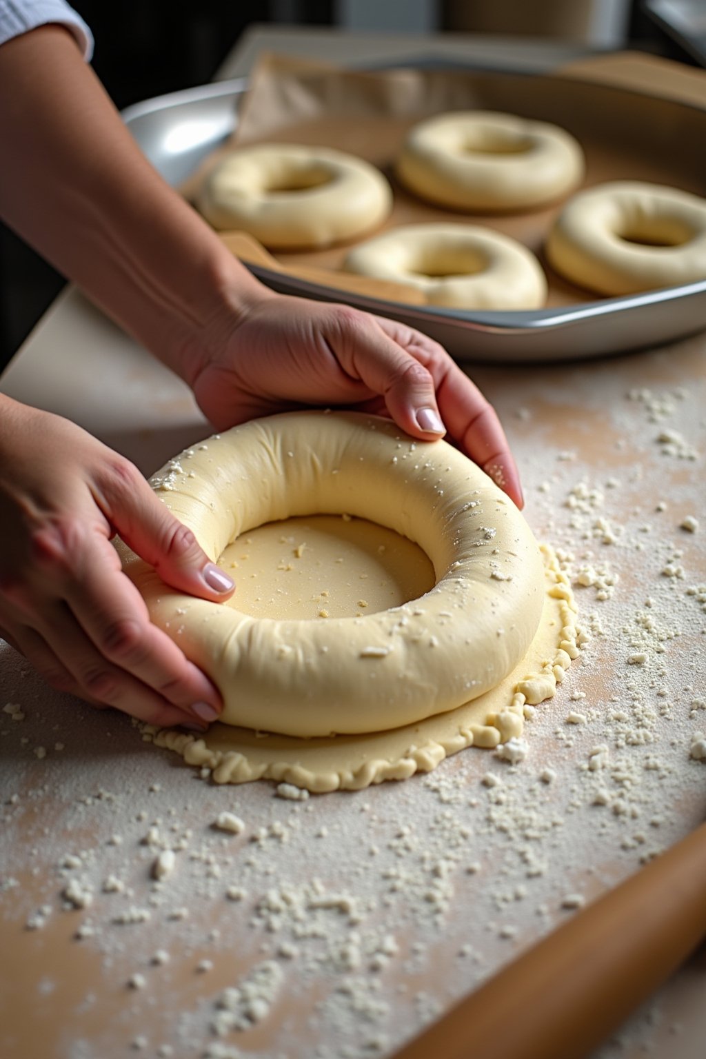 Hands shaping a cottage cheese bagel dough rope into a circle on a lightly floured surface, other formed bagels visible on parchment-lined baking sheet nearby, dusting of flour on the work surface,...