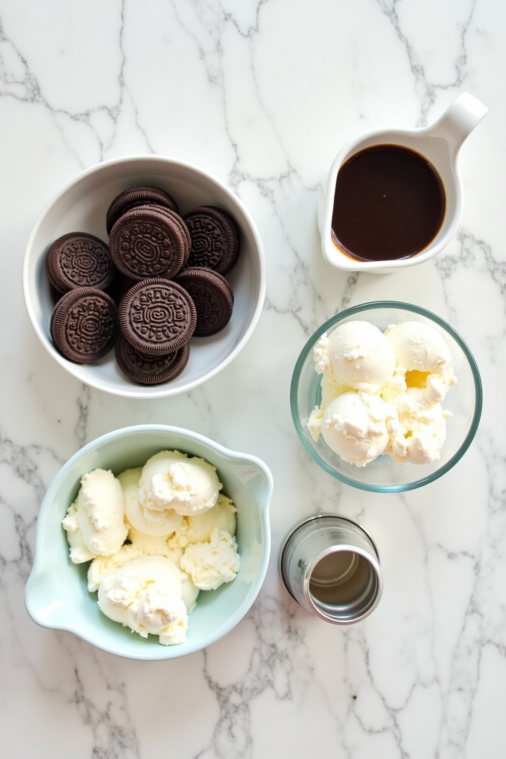 Flat lay mise en place of Oreo milkshake ingredients on a marble countertop, stack of Oreo cookies, bowl of vanilla ice cream scoops, small pitcher of milk, whipped cream canister, bottle of chocol...