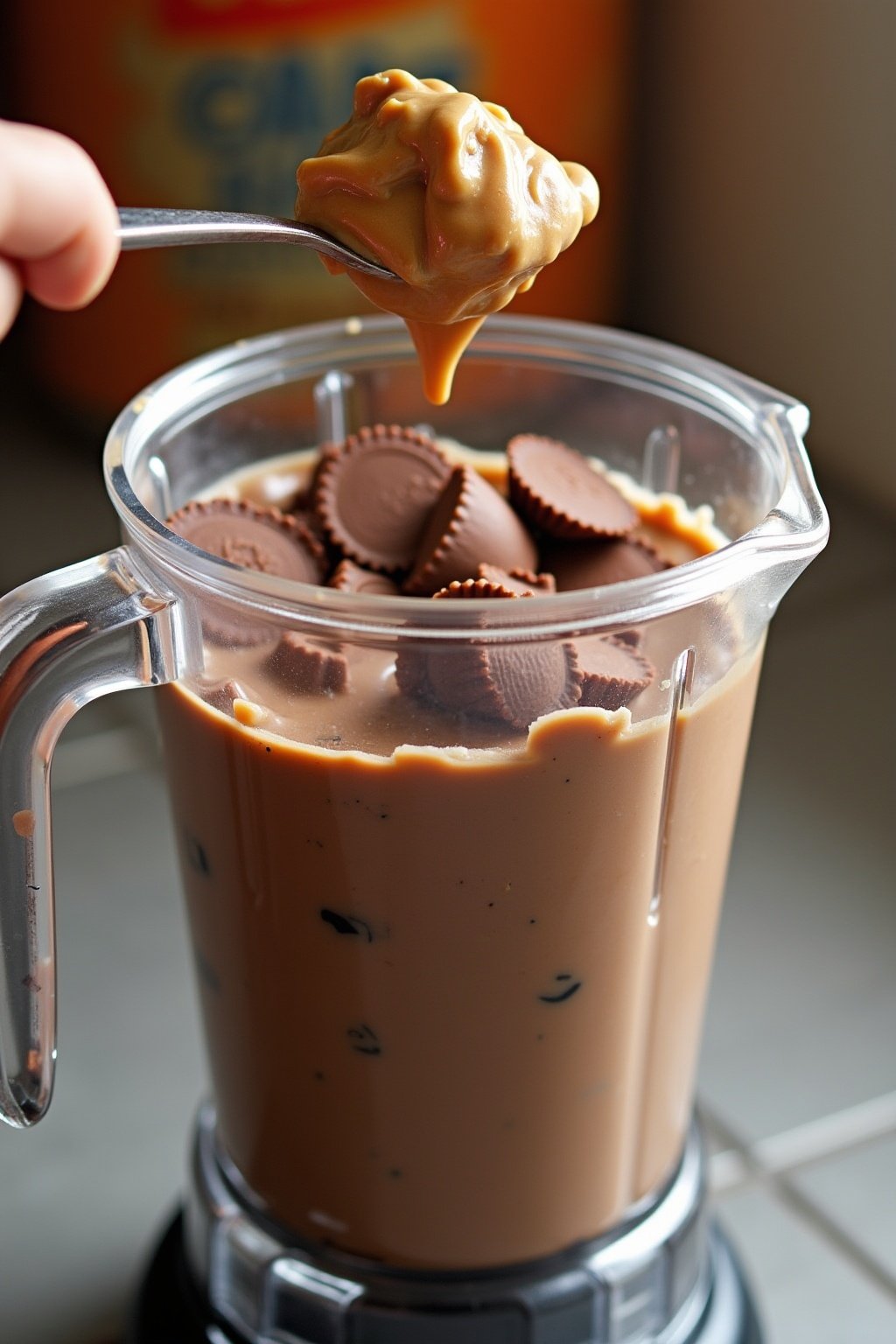Chopped Reeses peanut butter cups and a spoonful of peanut butter being dropped into a blender with chocolate ice cream, peanut butter swirling into dark chocolate mixture, creamy texture visible, ...