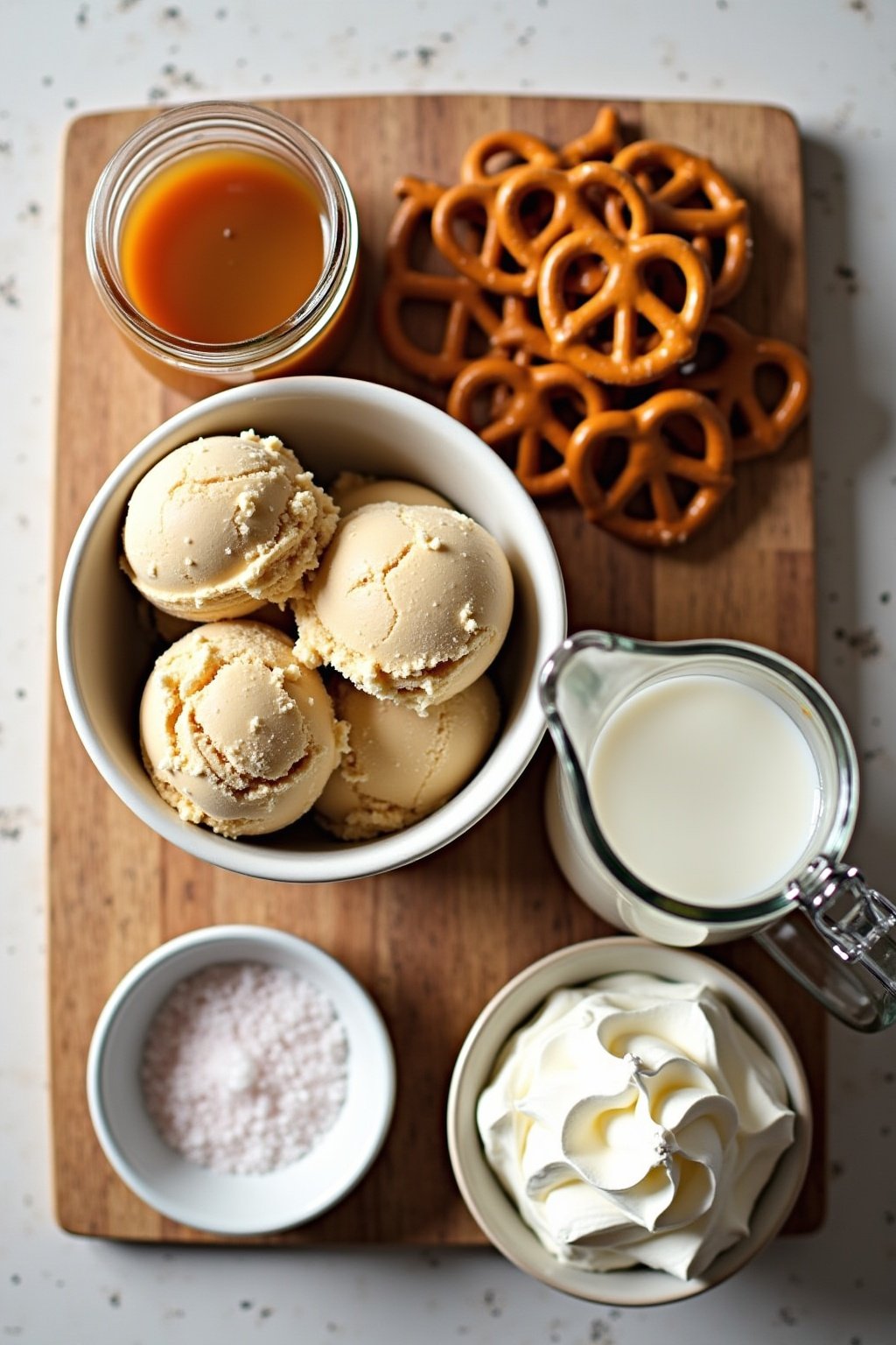 Flat lay of salted caramel pretzel milkshake ingredients on a wooden cutting board, bowl of caramel ice cream scoops, pile of mini pretzels, jar of salted caramel sauce, small pitcher of milk, flak...